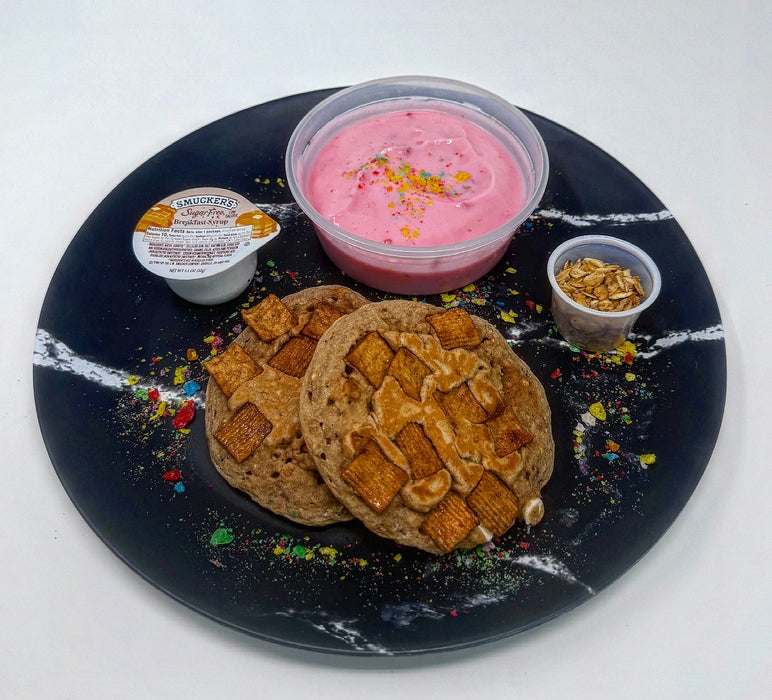 Two cookies with a pink dessert and small container on a black plate with white rim. The halal meal preps.
