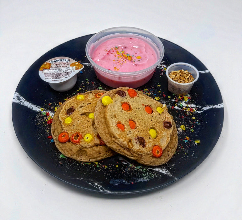 Stack of pancakes with colorful toppings on a black plate with side dishes including a pink yogurt and granola. A meal made by the halal meal prep.