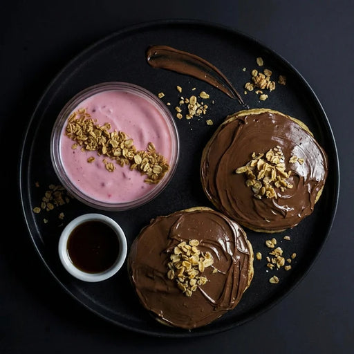 Two chocolate-glazed donuts with a side of pink yogurt and granola on a black plate.