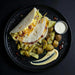 Plated dish with tortilla wrap, potatoes, and a side of sauce on a dark surface