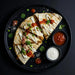 Quesadillas on a black plate with condiments on a dark background