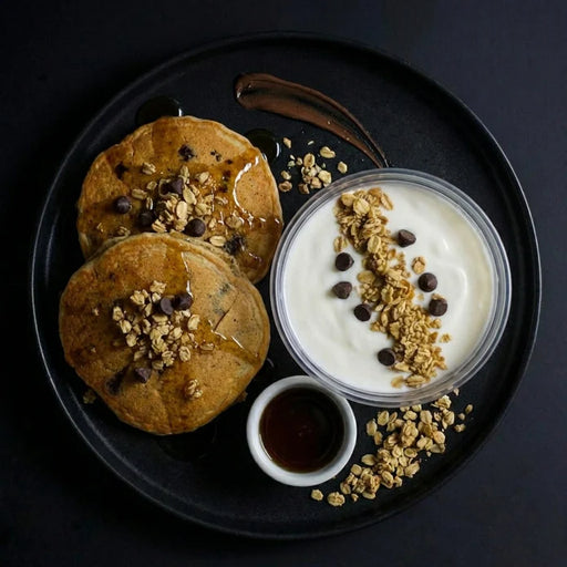 Two pancakes with syrup, granola, and chocolate chips on a black plate with yogurt and honey.