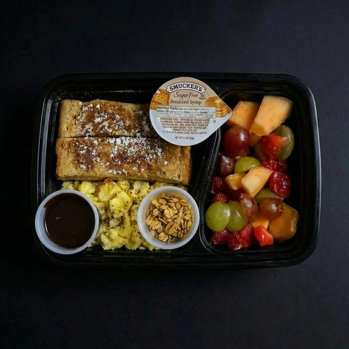 Breakfast meal in a black tray with French toast, fruit, and scrambled eggs on a dark background.