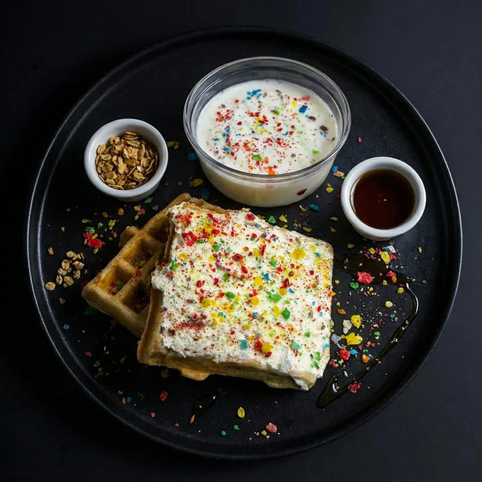 Waffle with colorful toppings on a black plate with syrup and granola.