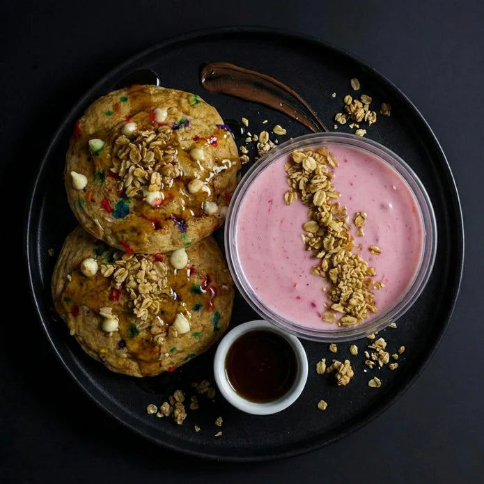 Two cookies with sprinkles and a pink yogurt bowl with granola on a black plate.